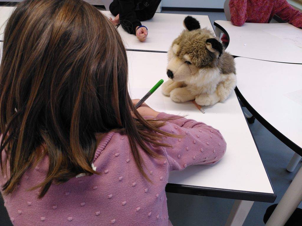 Giraffensprache ein kind mit braunen haaren, das einen rosa pullover trägt, schreibt an einem weißen tisch vor einem plüschwolf. andere kinder sitzen in der nähe, teilweise sichtbar, und arbeiten ebenfalls an den tischen.