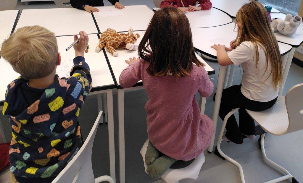 Giraffensprache vier kleine kinder sitzen an einem weißen tisch und zeichnen auf papier. zwei plüschtiere, eine giraffe und ein elefant, liegen auf dem tisch. die kinder werden von hinten betrachtet und scheinen auf ihre tätigkeit konzentriert zu sein.