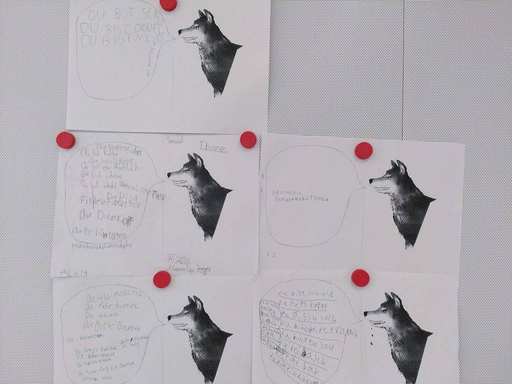 Giraffensprache sechs schwarz weiße arbeitsblätter mit wolfsillustrationen sind an eine tafel gepinnt. jedes blatt enthält handgeschriebenen deutschen text in und um sprechblasen, die an den wolf gerichtet sind. vier rote stecknadeln halten die blätter an ihrem platz.