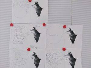 sechs schwarz weiße arbeitsblätter mit wolfsillustrationen sind an eine tafel gepinnt. jedes blatt enthält handgeschriebenen deutschen text in und um sprechblasen, die an den wolf gerichtet sind. vier rote stecknadeln halten die blätter an ihrem platz.