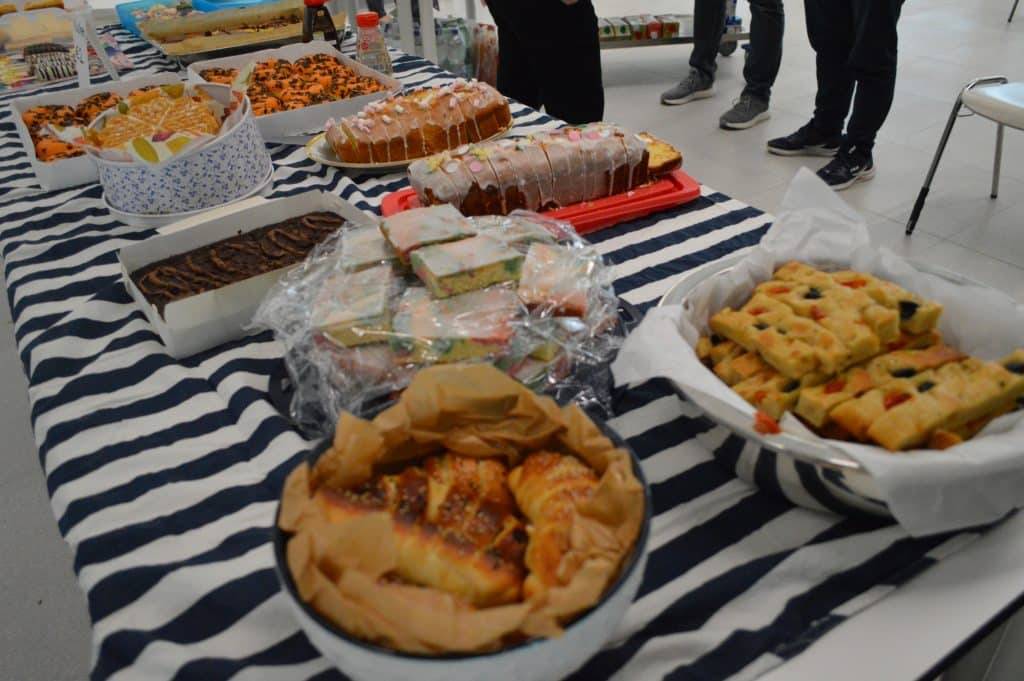 ein mit einem gestreiften tuch bedeckter tisch ist mit verschiedenen backwaren, darunter kuchen, focaccia, gebäck und sandwiches, belegt. mehrere personen stehen um den tisch herum, ihre beine sind im hintergrund zu sehen. ein mit einem gestreiften tuch bedeckter tisch ist mit verschiedenen backwaren, darunter kuchen, focaccia, gebäck und sandwiches, belegt. mehrere personen stehen um den tisch herum, ihre beine sind im hintergrund zu sehen.