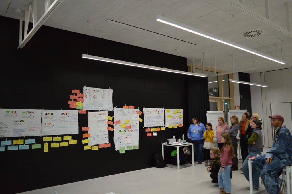 eine gruppe von menschen, darunter kinder und erwachsene, steht und sitzt vor einer schwarzen wand, die mit bunten postern und haftnotizen in einem modernen, gut beleuchteten raum bedeckt ist. eine gruppe von menschen, darunter kinder und erwachsene, steht und sitzt vor einer schwarzen wand, die mit bunten postern und haftnotizen in einem modernen, gut beleuchteten raum bedeckt ist.
