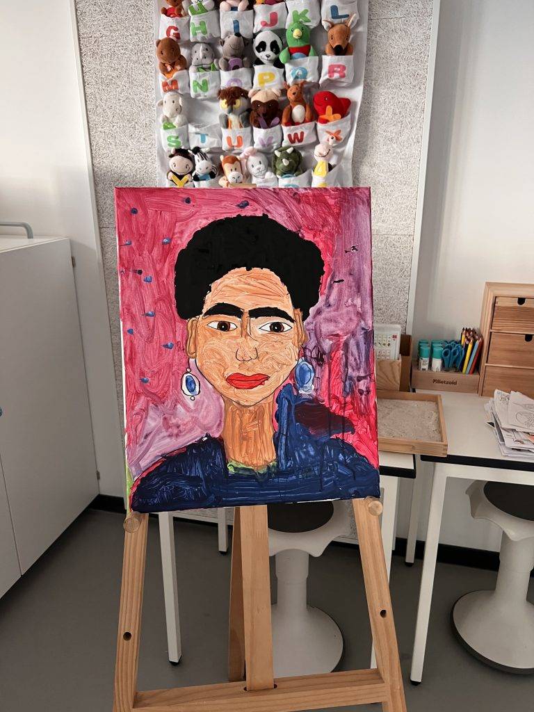 ein farbenfrohes gemälde einer frau mit ohrringen, dunklem haar und einem blauen oberteil ist auf einer staffelei in einem raum mit spielzeug, kunstzubehör und kindermöbeln ausgestellt. der hintergrund des bildes ist rosa.