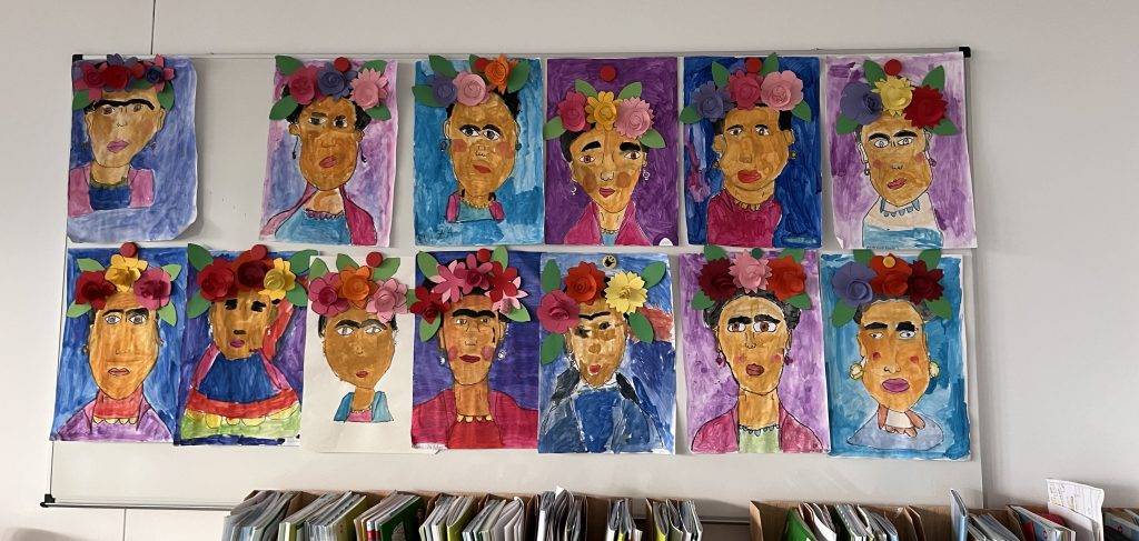 an einer wand im klassenzimmer hängen farbenfrohe porträts von frida kahlo, die sich durch helle hintergründe, markante gesichtszüge und 3d papierblumen als kopfschmuck auszeichnen. in einem regal unterhalb der kunstwerke sind reihen von büchern zu sehen.