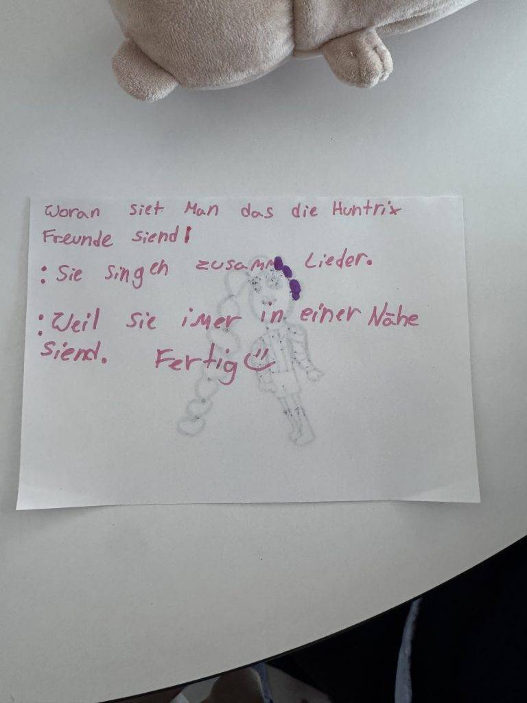 KPop Demon Hunters eine handschriftliche notiz in deutscher sprache auf weißem papier, verziert mit einer schwachen skizze von zwei personen. am oberen und unteren rand des bildes sind ein plüschtier und ein teil eines runden tisches zu sehen.