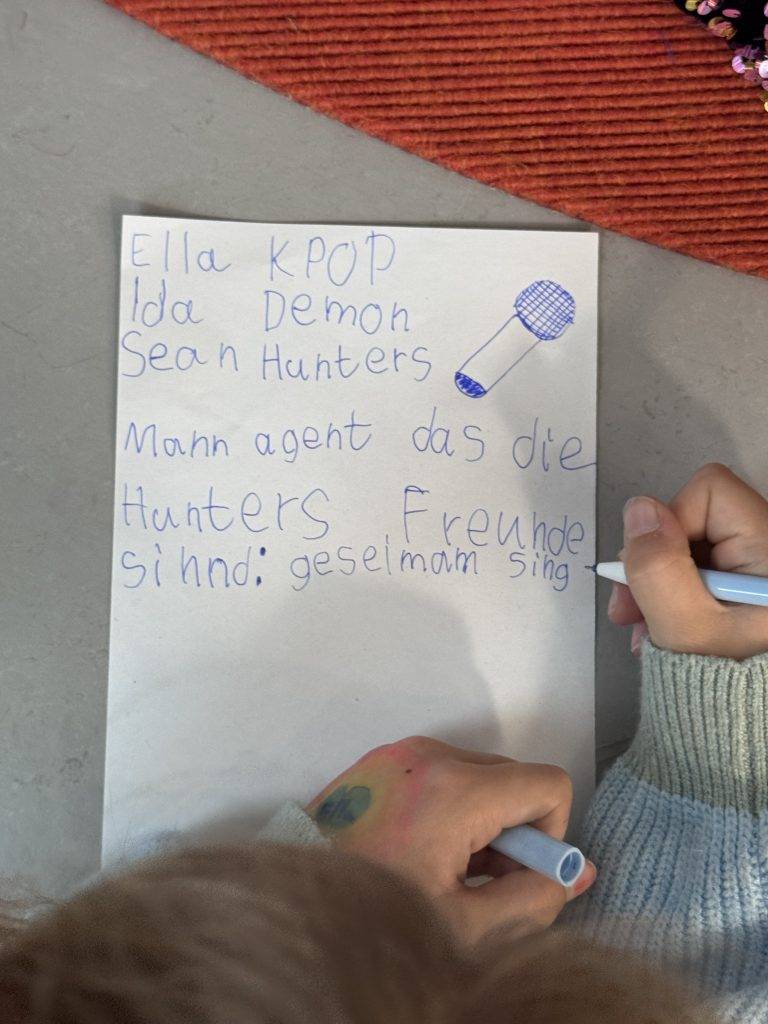 KPop Demon Hunters ein kind schreibt mit blauem filzstift auf ein blatt papier. auf dem zettel, der in deutscher sprache verfasst ist, stehen namen, ein kritzelndes mikrofon und die worte freunde sind: gemeinsam singen. man sieht die hände des kindes, die den marker halten.
