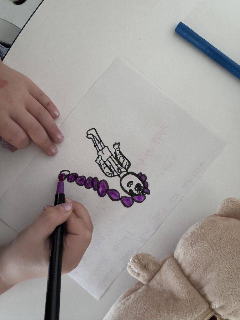 KPop Demon Hunters ein kind malt die zeichnung einer figur auf einem weißen blatt papier mit einem lila marker aus. ein weiterer blauer marker und ein plüschbär liegen in der nähe auf dem weißen tisch.