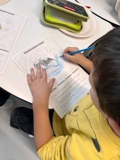 Unser Wasser-Projekt ein kind in einem gelben pikachu shirt malt mit einem blauen farbstift ein arbeitsblatt mit dem titel "winter animal scene" an einem tisch aus. ein grünes tablett und ein weiteres arbeitsblatt liegen ebenfalls auf dem tisch.