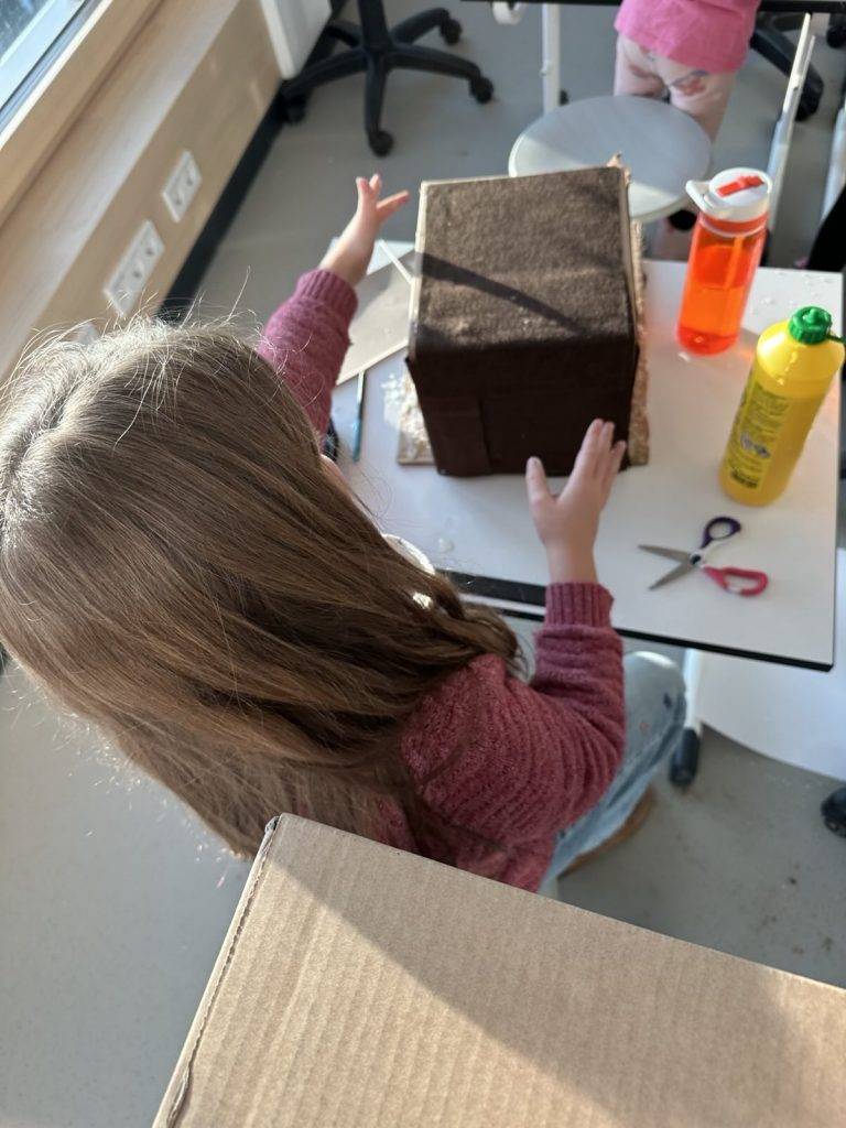 Miniaturwelten ein kind mit langen braunen haaren und einem rosa pullover sitzt an einem tisch und arbeitet an einem braunen karton. auf dem tisch liegen eine schere, eine wasserflasche und eine gelbe flasche. das sonnenlicht fällt durch das fenster.