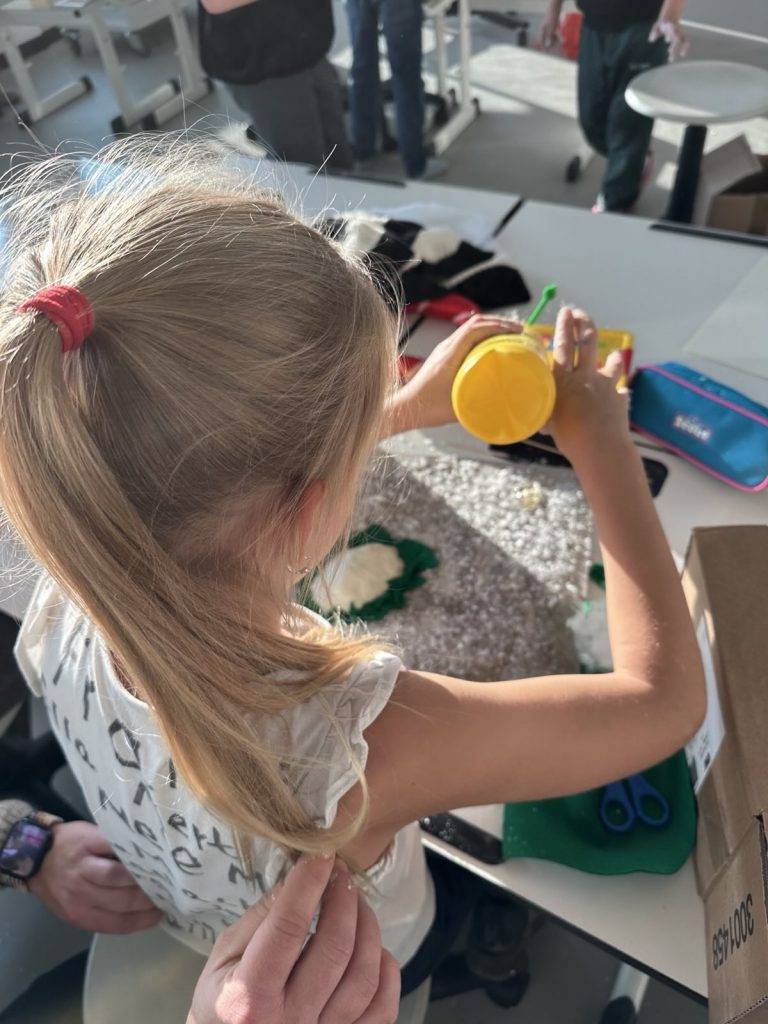 Miniaturwelten ein junges mädchen mit blondem haar in einem pferdeschwanz arbeitet an einem bastelprojekt an einem tisch und schüttet gelbes pulver auf eine strukturierte oberfläche, umgeben von bastelmaterial und anderen schülern in einem hell erleuchteten klassenzimmer.