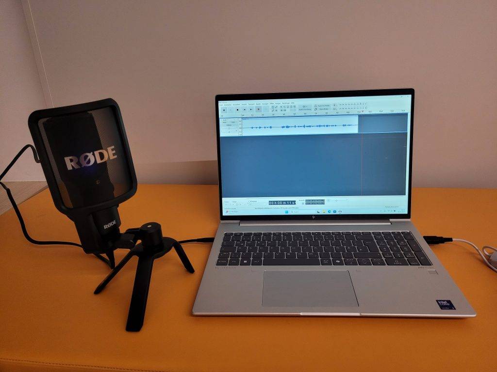 ein rØde mikrofon auf einem kleinen stativ ist mit einem silbernen laptop verbunden, auf dessen bildschirm eine software zur audiobearbeitung mit einer wellenform angezeigt wird. beide sind auf einer gelben fläche vor einer schlichten wand platziert.