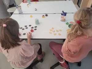 zwei kinder sitzen an einem tisch und ordnen kleine, bunte keksformen an. ausstechformen und schachteln liegen ebenfalls auf dem tisch. die kinder werden von hinten betrachtet, während sie mit den keksen und backutensilien arbeiten.