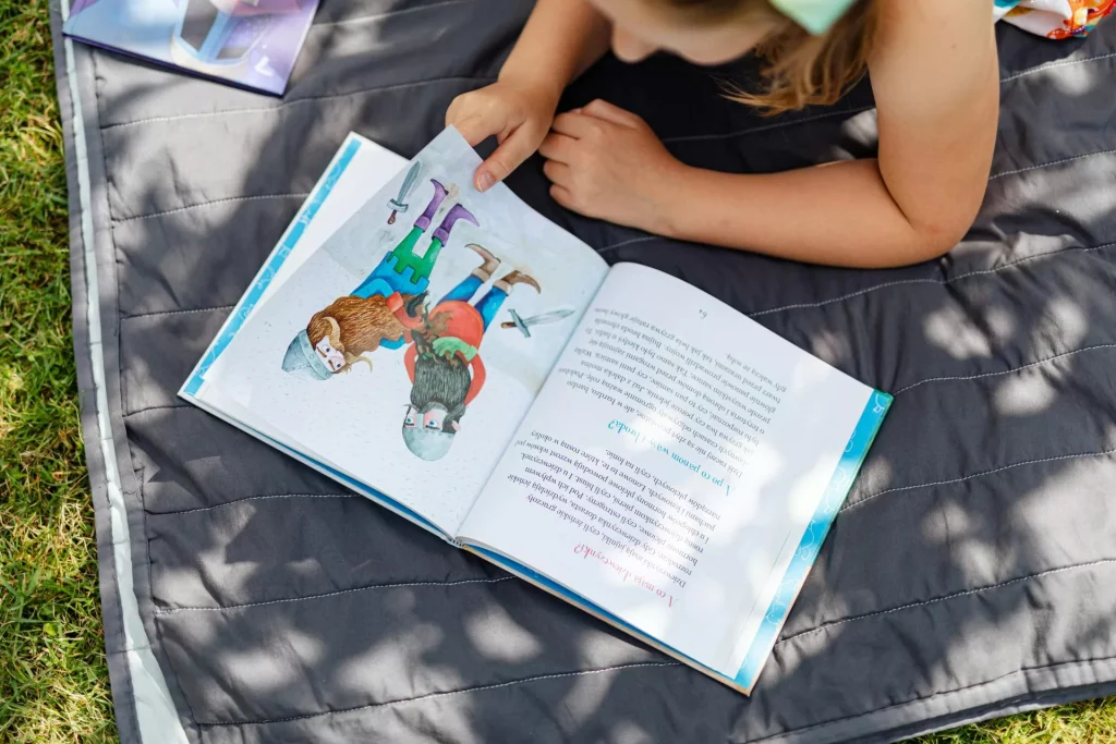 ein kind liegt auf einer grauen decke im freien und liest ein illustriertes buch, das zwei mädchen mit tieren zeigt. das sonnenlicht fällt durch die Äste der bäume und wirft schatten auf die seite und die decke. ein kind liegt auf einer grauen decke im freien und liest ein illustriertes buch, das zwei mädchen mit tieren zeigt. das sonnenlicht fällt durch die Äste der bäume und wirft schatten auf die seite und die decke.
