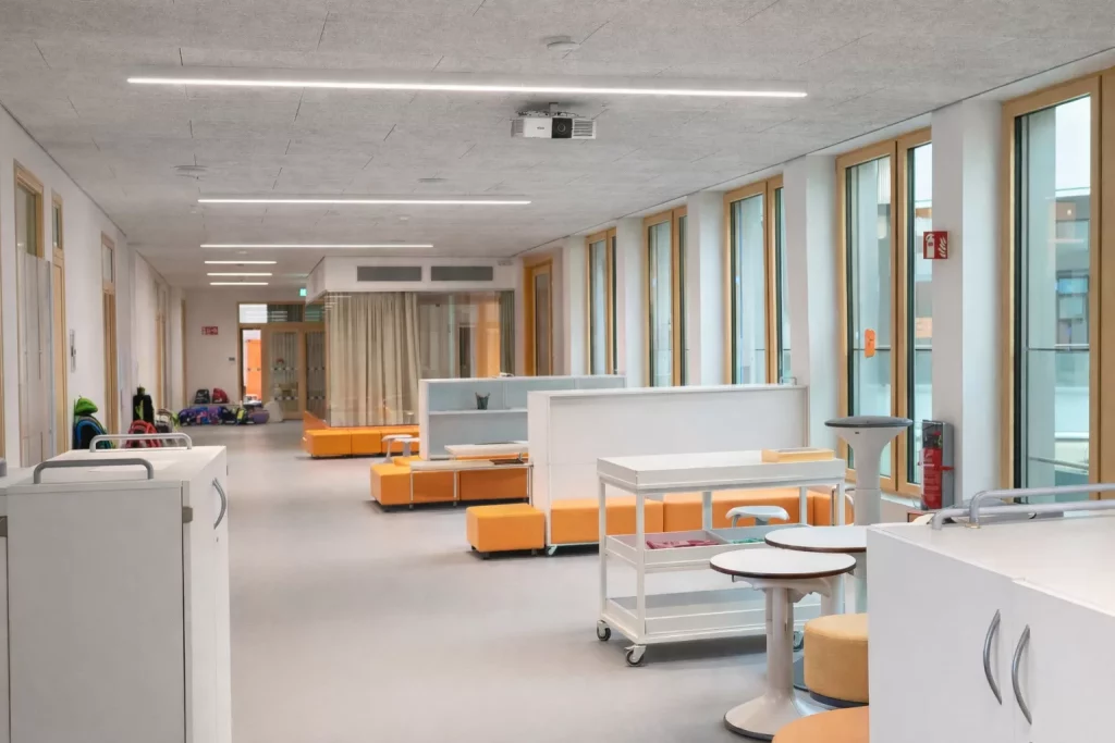 ein moderner, geräumiger krankenhausflur mit großen fenstern, orangefarbenen sitzgelegenheiten, weißen trennwänden und medizinischen wagen. durch die fenster strömt licht herein und schafft eine helle und saubere atmosphäre.