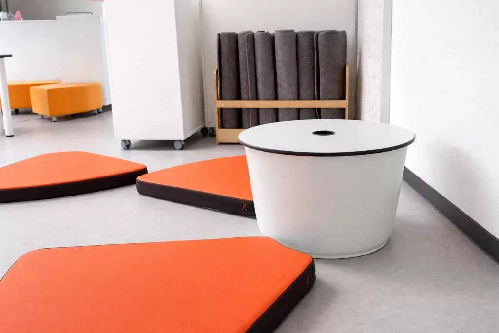 ein moderner raum mit drei orangefarbenen bodenkissen, einem runden weißen tisch und einem regal mit mehreren aufgerollten dunkelgrauen matten vor einer hellen wand.