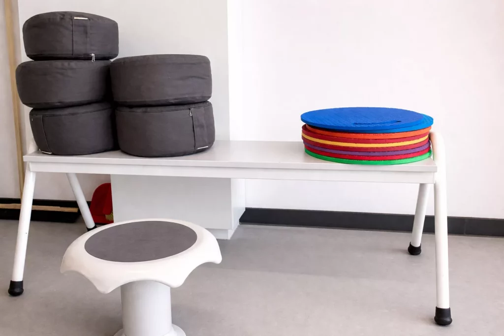 auf einem weißen tisch liegen vier gestapelte graue kissen auf der linken seite und fünf bunte runde matten auf der rechten seite. vor dem tisch steht ein weißer hocker mit einer grauen sitzfläche. die wand im hintergrund ist schlicht und weiß.