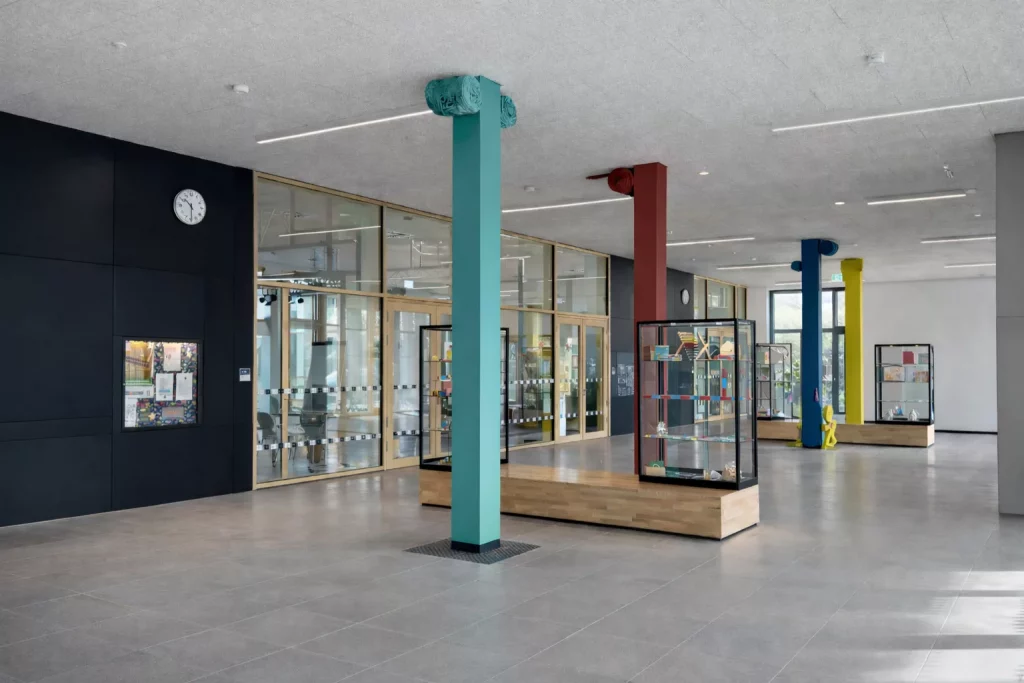 ein geräumiger, moderner schulflur mit bunt bemalten säulen, glasvitrinen, in denen schülerarbeiten ausgestellt sind, großen fenstern und einer uhr an einer schwarzen wand. der raum ist hell und offen und hat einen grauen bodenbelag.