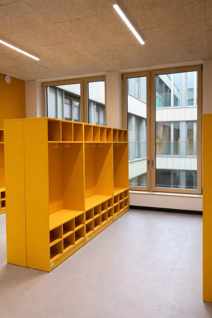 leuchtend gelbe fächerschränke mit regalen und haken sind in einem modernen klassenzimmer oder kindertagesstättenraum mit großen fenstern und viel natürlichem licht in reihen angeordnet.
