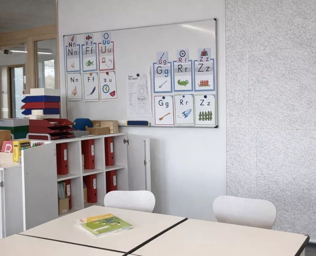ein klassenzimmer mit weißen tischen und stühlen, einem bücherregal mit roten ordnern und einer tafel mit bunten buchstabentafeln und illustrationen. ein kinderbuch liegt aufgeschlagen auf dem tisch.