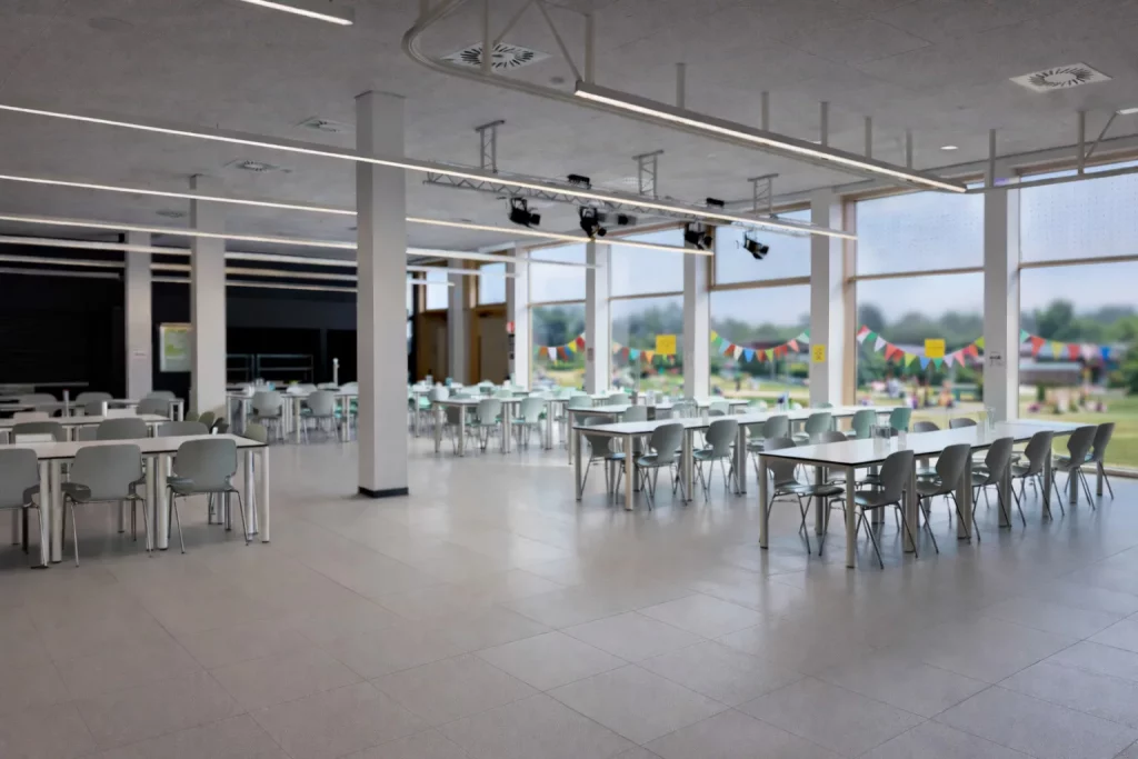 eine geräumige, moderne cafeteria mit reihen leerer weißer tische und stühle, großen fenstern, die natürliches licht hereinlassen, und bunten wimpeln, die draußen auf einer wiese zu sehen sind.
