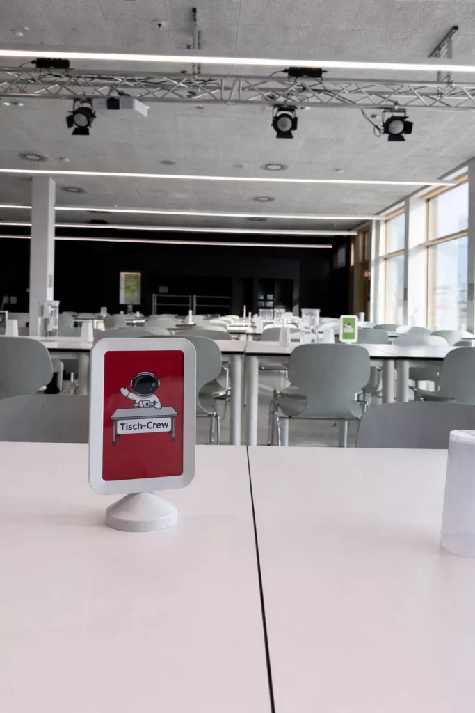 ein weißer tisch in einer modernen, leeren cafeteria mit reihen von grauen stühlen. ein kleines schild auf dem tisch trägt die aufschrift tisch crew und zeigt die abbildung einer person mit einem tablett. große fenster lassen natürliches licht herein.