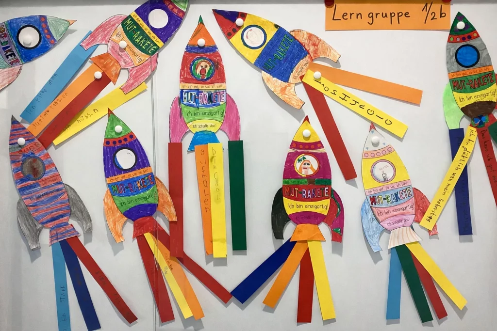 bunte papierraketen mit handgeschriebenen botschaften und fotos von kindern sind an einer weißen wand unter einem schild mit der aufschrift "lern gruppe 1/2b" angebracht. jede rakete hat lange, bunte papierschlangen als flammen.