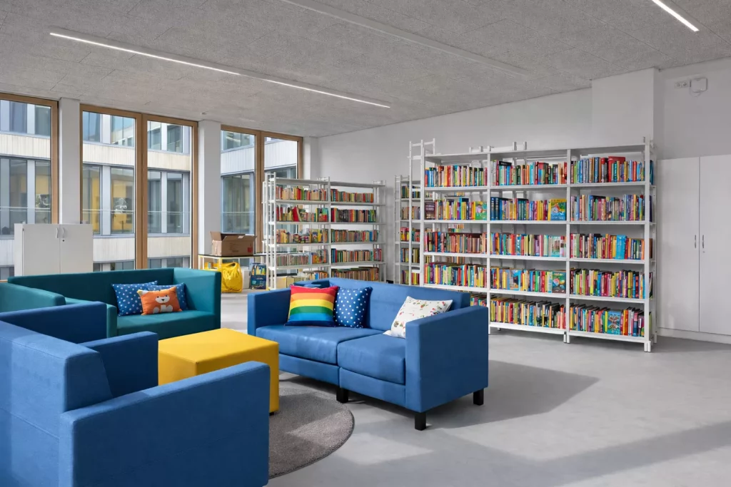 eine moderne bibliothek mit blauen sofas, bunten kissen, einem gelben tisch und mit büchern gefüllten bücherregalen. große fenster lassen natürliches licht herein, und der raum wirkt sauber und einladend.