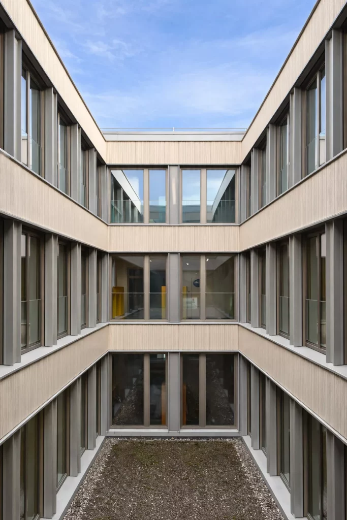 ein modernes, dreistöckiges gebäude mit großen fenstern und einem zentralen, offenen, mit kies gefüllten innenhof, der zum blauen himmel hinaufragt. die fassade ist beige mit vertikalen metallakzenten.