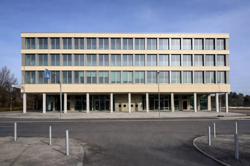 ein modernes, rechteckiges, vierstöckiges gebäude mit großen glasfenstern, beigem Äußeren und einem überdachten, von säulen getragenen eingang. der platz davor ist gepflastert und mit einigen straßenschildern und pollern versehen. der himmel ist klar.