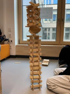 ein hoher, verschlungener turm aus holzbrettern steht in einem innenraum in der nähe eines fensters, umgeben von menschen, die auf dem boden sitzen. der turm weist gestaffelte und ineinandergreifende schichten auf, mit einer runden struktur nahe der spitze.