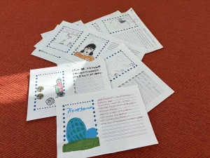 mehrere illustrierte und handgeschriebene arbeitsblätter für kinder sind auf einer orangefarbenen, strukturierten oberfläche ausgebreitet, mit zeichnungen, gedruckten gepunkteten rändern und linierten feldern, die mit schrift gefüllt sind.