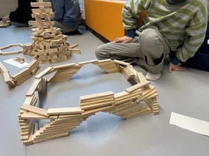 ein kind in einem grün gestreiften pullover sitzt auf dem boden in der nähe von holzklötzchenstrukturen, darunter ein rundbogen und ein turm mit der aufschrift kapla. im hintergrund sind weitere kinder und klötze zu sehen.