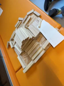 auf einer orangefarbenen bank steht ein holzblock, auf dem ein stück papier mit der aufschrift emmale grant liegt. im hintergrund sind die füße einer person in turnschuhen zu sehen.