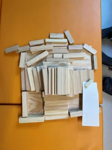 auf einer orangefarbenen fläche ist eine gruppe von holzklötzen angeordnet, die einer struktur oder einem gebäude ähneln. ein weißes stück papier mit handgeschriebenem text befindet sich in der unteren rechten ecke, teilweise unter den blöcken.
