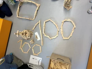 auf einem grauen boden sind bauklötze aus holz in verschiedenen geometrischen formen angeordnet, darunter polygone und ein hohes gebäude in der mitte. kinder sitzen in der nähe, und eine kiste mit zusätzlichen bauklötzen ist sichtbar.