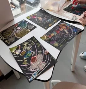 vier bunte buntstift und glitzerzeichnungen des sonnensystems auf schwarzem papier sind auf einem kleinen weißen tisch in der nähe eines fensters ausgestellt. in der oberen rechten ecke ist eine hand mit bastelmaterial zu sehen.