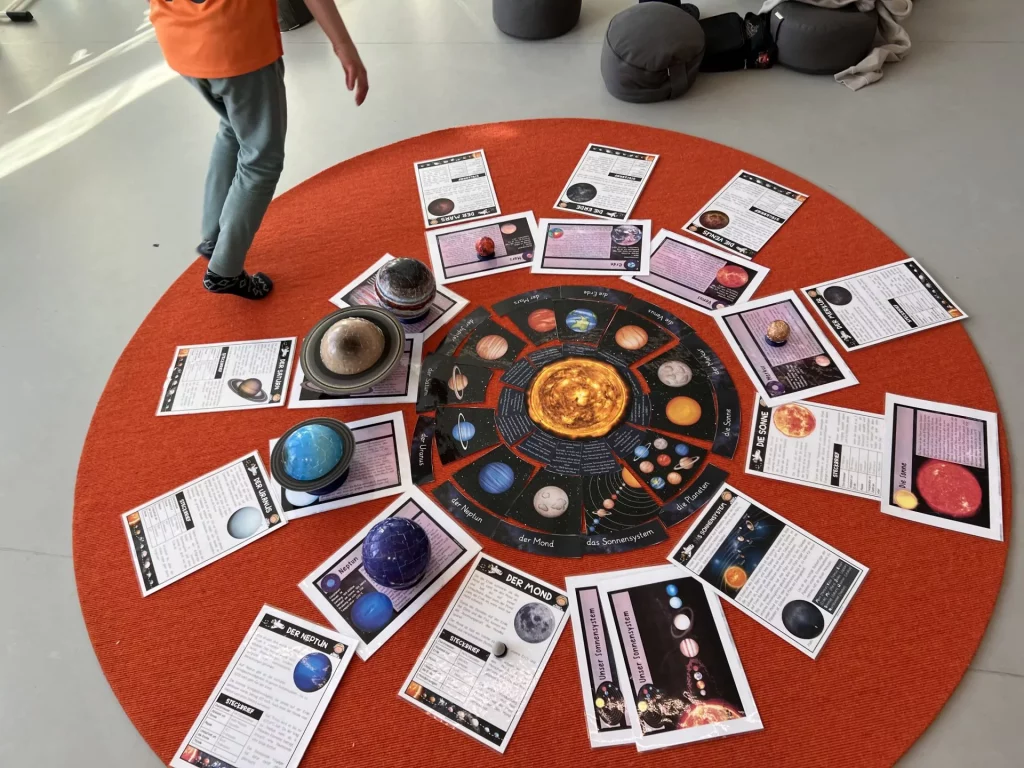 ein kreisförmiges display auf einem orangefarbenen teppich zeigt ein modell des sonnensystems mit 3d planetenmodellen und faktenblättern, die um eine zentrale sonne angeordnet sind. eine person geht in der nähe spazieren.