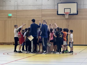 eine gruppe von kindern und zwei erwachsenen steht in einer turnhalle unter einem basketballkorb und drückt sich an den händen zu einem team zusammen. einige kinder tragen sporttrikots, ein erwachsener hält ein klemmbrett in der hand. die turnhalle hat holzwände und einen gelben boden.