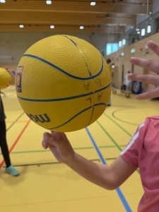 eine person in einem rosa hemd dreht einen gelben basketball auf ihrem finger in einer sporthalle mit gelbem boden und bunten linien. eine andere person steht im hintergrund.