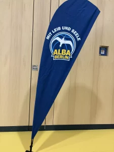 ein blaues, tropfenförmiges banner mit dem text mit leib und seele und alba berlin unter einem weißen, fliegenden vogel logo, das in einem innenraum vor einer hellen holzwand steht.