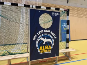 ein schild mit dem logo der basketballmannschaft alba berlin und der aufschrift "mit leib und seele" steht vor einem tor in einer sporthalle. im hintergrund sind sportnetze, eine bank und eine holzwand zu sehen.