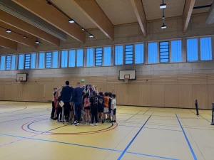 eine gruppe von kindern und zwei erwachsene stehen mit erhobenen armen in einem kreis in einer turnhalle mit hohen fenstern, holzwänden und basketballkörben.